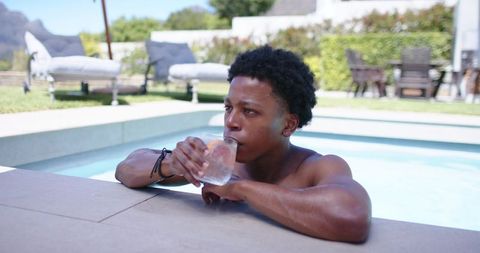 Shirtless man sipping cold drink while resting on pool edge, relaxing in sunny backyard
