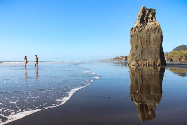 Two people admiring large rock formation on vast beach with clear blue sky, ocean waves gently hitting shore. Ideal for travel promotion, leisure activities, or nature exploration themes.