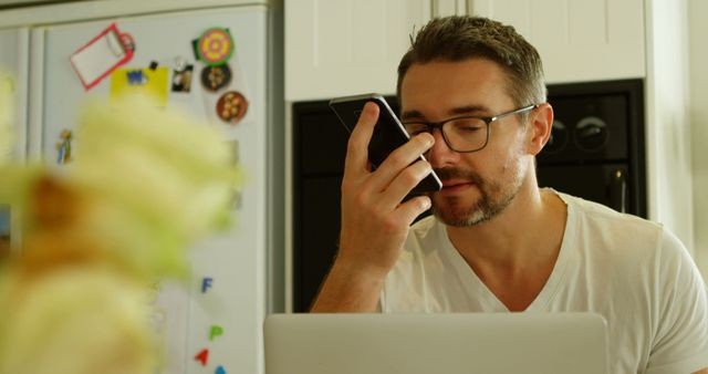 Young man sitting at kitchen table using smartphone voice command while working on laptop. Wears glasses and casual clothing, indicating a modern work-from-home setup. Ideal for use in articles or advertisements related to remote work, technology, modern work culture, and lifestyle.