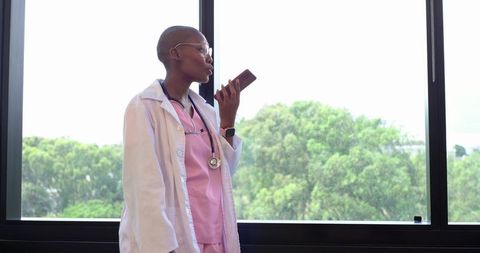African clinician speaking into smartphone by window wearing lab coat and pink scrubs