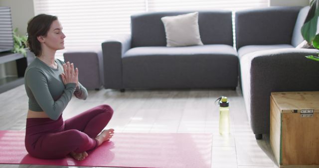 Tranquil scene of a young woman practicing yoga and meditation at home seated on a pink mat with a water bottle nearby. Ideal for wellness blogs, fitness advertisements, and home workout instructional content.
