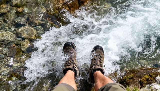 Hiker soaking boots in crystal mountain stream rapids with mossy rocks and sunlit foam