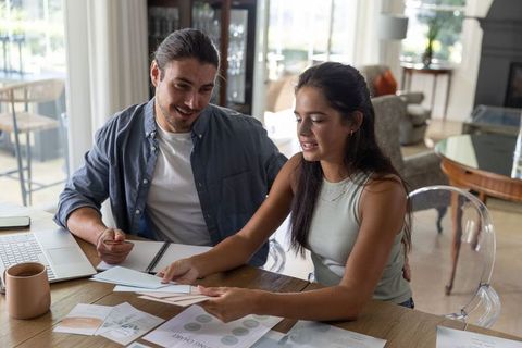 Couple reviewing budget and home projects at wooden table with laptop, envelopes, charts