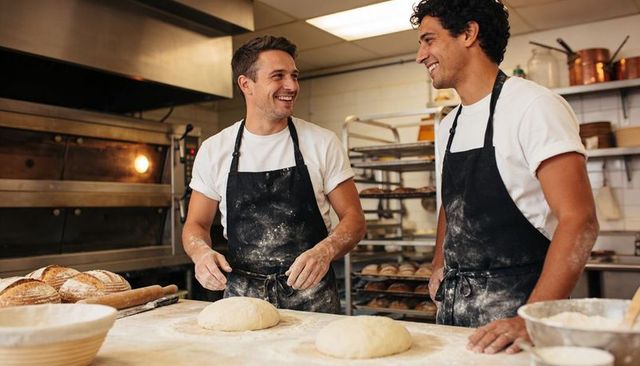 Two bakers kneading dough in busy bakery kitchen smiling teamwork artisan breadmaking