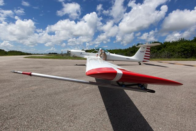 Motorized glider parked on runway at Kennedy Space Center ready for take-off to measure sonic booms as part of the SonicBAT II Program. F-18 jets from NASA flying at supersonic speeds generate the booms to study their effects in low-altitude turbulence. Possible uses include articles on aviation technology, scientific studies on sonic booms, aerospace research, NASA programs, and educational materials.