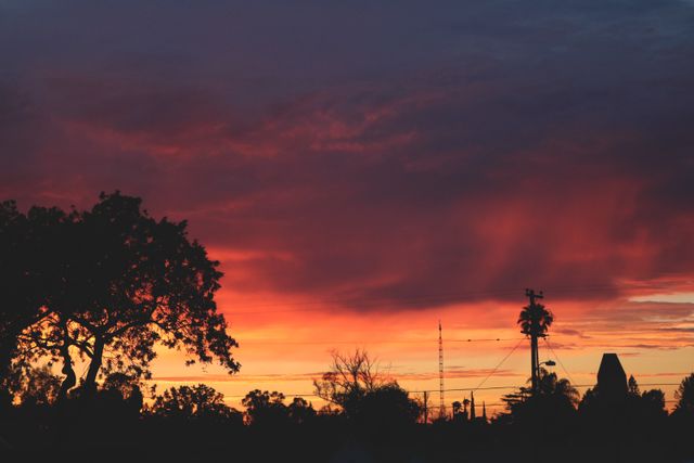 This image showcases a vibrant sunset over an urban landscape with silhouetted trees and structures. The fiery colors of the sunset contrast beautifully with the dark foreground, creating a stunning visual effect. Ideal for use in nature and travel blogs, background images, web design, and promotional materials emphasizing urban beauty and natural splendor.