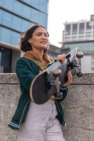 Young woman carrying skateboard in urban plaza wearing green jacket and brown scarf