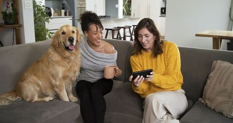 Friends sharing tablet and coffee on couch with golden retriever in cozy open-plan living room