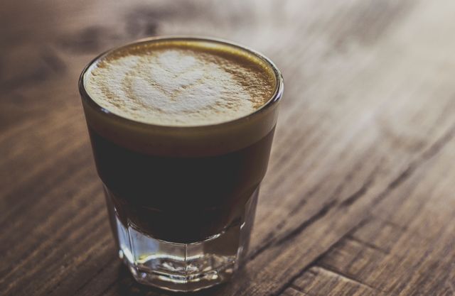 Close-up photograph of a freshly brewed cortado coffee served in a clear glass mug placed on a rustic wooden table. The cortado features intricate latte art in the foam, adding to its visual appeal. This image is perfect for use in coffee shop promotions, breakfast menu designs, barista training manuals, or social media content for cafes and coffee enthusiasts.