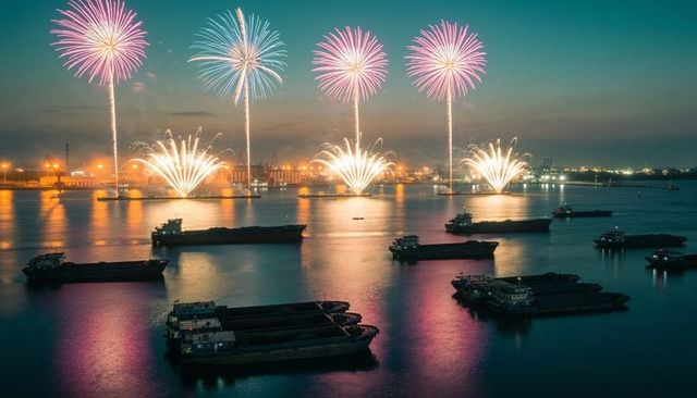 Harbor fireworks display over cargo barges reflecting neon fountains and port lights