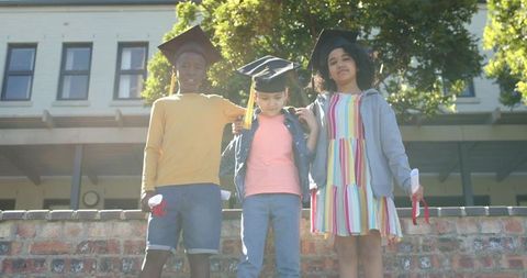 Children celebrating graduation standing on brick wall at school holding diplomas and smiling