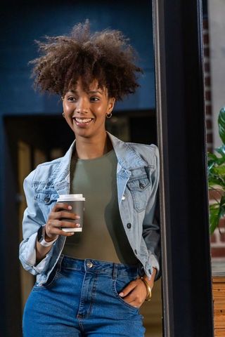 Smiling african woman holding takeaway coffee in denim jacket at cafe entrance