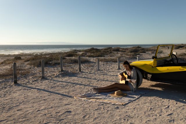 Man sitting on sandy beach near yellow beach buggy, playing guitar at sunset. Ideal for themes of relaxation, summer vacations, road trips, and outdoor adventures. Perfect for travel blogs, music-related content, and lifestyle promotions.