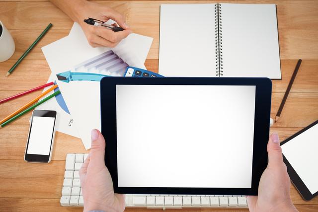 Hands using a digital tablet with blank screen, placed on office desk filled with stationery including notebook, pens, and smartphone. Ideal for visuals related to remote work, office environment, productivity, and digital technology.
