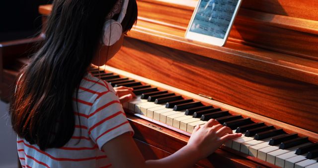 Young girl wearing headphones playing piano while watching lesson on tablet. Perfect for themes related to childhood education, online learning, and music practice. Ideal for use in educational content, music tutorials, and parent-child learning activities.