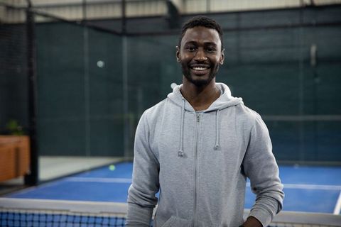 African american man smiling and posing on blue padel court wearing gray hoodie after warm-up