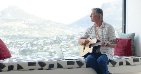 Contemplative man playing acoustic guitar on window bench with panoramic mountain view