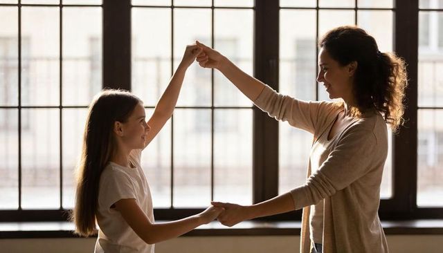 Mother and daughter dancing in sunlit room by large grid window, family bonding