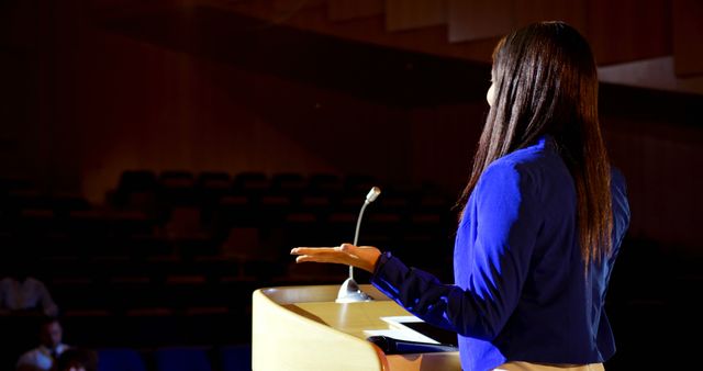 Woman confidently giving presentation on stage to an audience in a poorly lit auditorium. Ideal for content about public speaking, leadership skills, communication workshops, business seminars, and educational events.