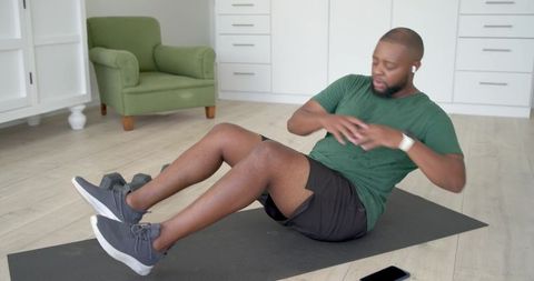 African american man performing seated russian twist at home on mat with dumbbells