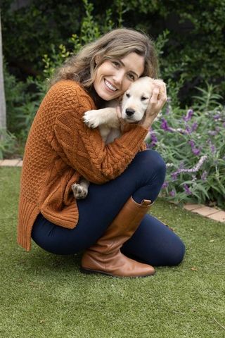 Smiling woman cuddling golden retriever puppy in garden wearing rust sweater and tan boots