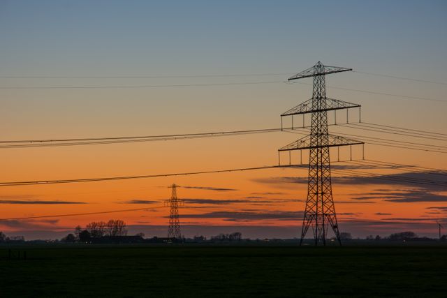 Electricity transmission towers are stretching across a rural landscape during a stunning sunset. The sky is painted with hues of orange and blue, with dark silhouettes of towers and trees visible in the distance. This visual could be used to depict themes of energy, infrastructure, and rural life, suitable for educational material, energy sectors, and environmental campaigns.