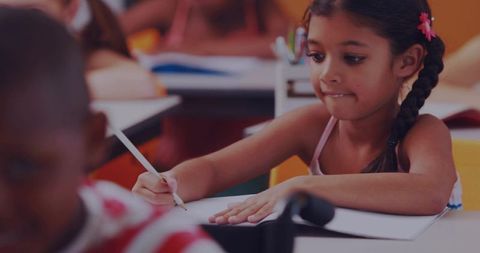 Young schoolgirl writing at desk in colorful classroom with braided hair and flower clip