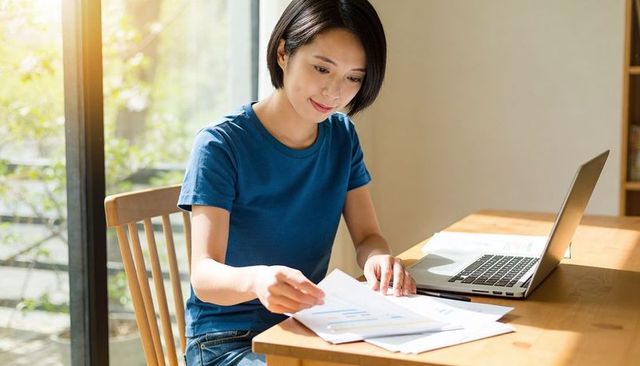 Asian woman working from home sorting financial charts by sunlit window with laptop