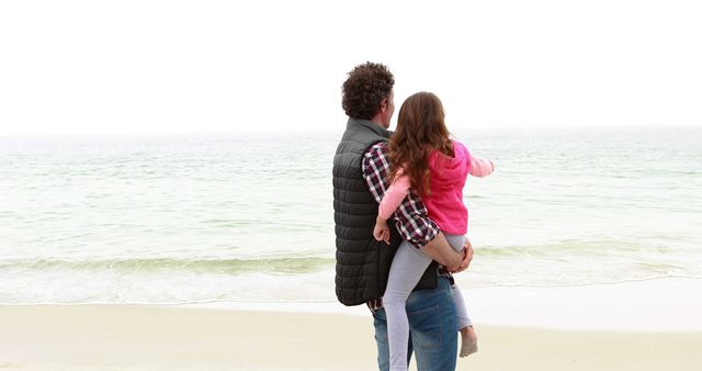 Father holding young daughter while standing on sandy beach, looking at ocean. Ideal for family, parenting, outdoor activities, vacation promotions, and father-daughter relationship themes.