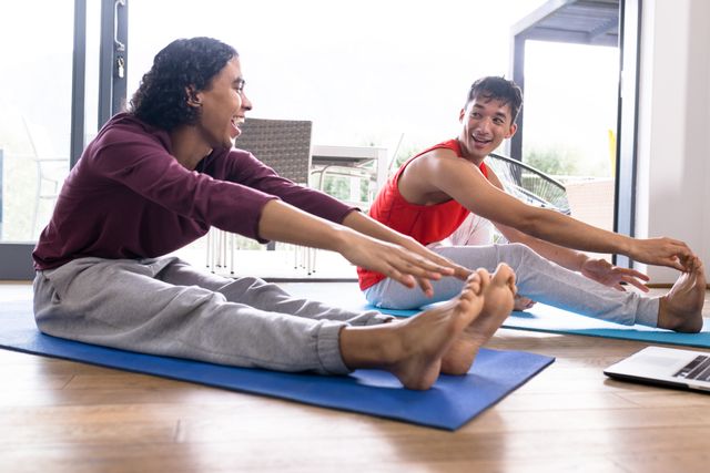 Multiracial gay couple practicing yoga together on mats at home, laughing and enjoying each other's company. This image is perfect for promoting healthy lifestyles, home fitness routines, and LGBTQ inclusivity. Ideal for use in wellness blogs, fitness apps, and social media campaigns focused on togetherness and love.