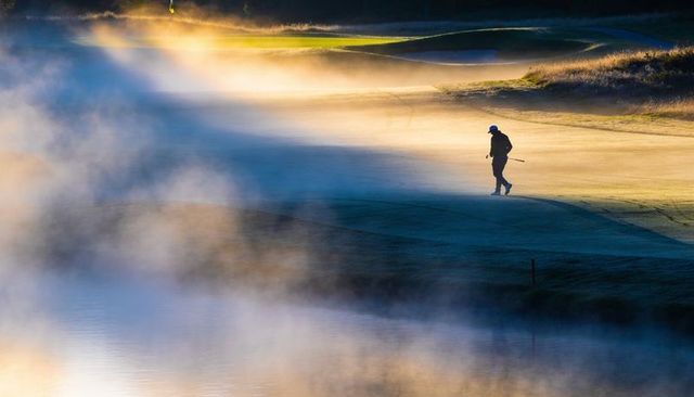 Solitary golfer walking misty sunrise fairway backlit silhouette reflective water