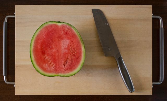 This stock photo displays a halved watermelon next to a stainless steel knife on a wooden cutting board. Ideal for use in food blogs, culinary articles, healthy eating promotions, kitchen-related content, and fruit cooking recipes. The image conveys freshness and simplicity, making it suitable for summer themes and healthy lifestyle visuals.