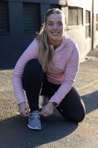Smiling young woman kneeling tying running shoe on sunlit urban pavement before workout