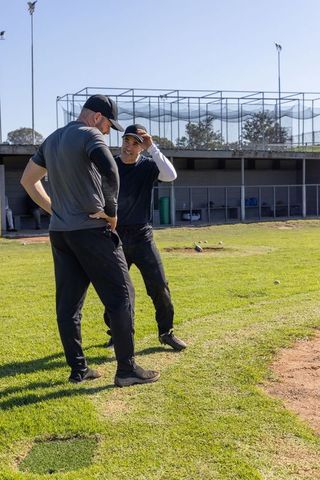 Baseball coach mentoring player on practice field adjusting cap near dugout sunny grass