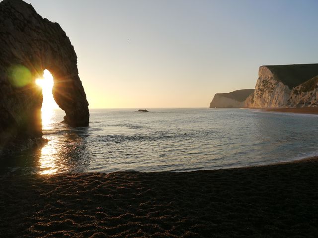 Beautiful sunset scene at Durdle Door, Dorset coastline. Shows the rock arch and sandy beach with the sun setting over the sea. Perfect for travel brochures, tourism advertisements, nature calendars, and desktop backgrounds.