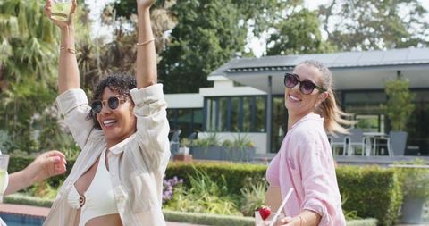 Friends celebrating poolside: diverse women wearing coverups and sunglasses with cocktails