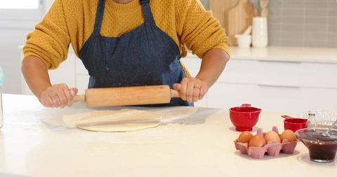 Home baker rolling dough on bright white countertop wearing mustard sweater and apron