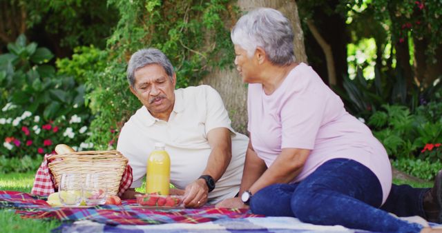 An elderly man and woman relaxing on a picnic blanket in a lush, green park. They are sharing fresh fruit and engaging in conversation, highlighting themes of companionship, healthy living, and outdoor activity. Ideal for use in health and wellness promotions, retirement community brochures, relationship advice blogs, and nature outing advertisements.