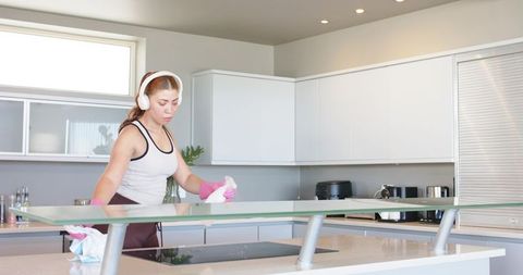 Woman cleaning modern kitchen island wearing pink gloves and headphones