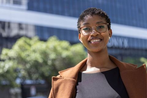 Confident young professional smiling in urban plaza wearing brown coat and glasses