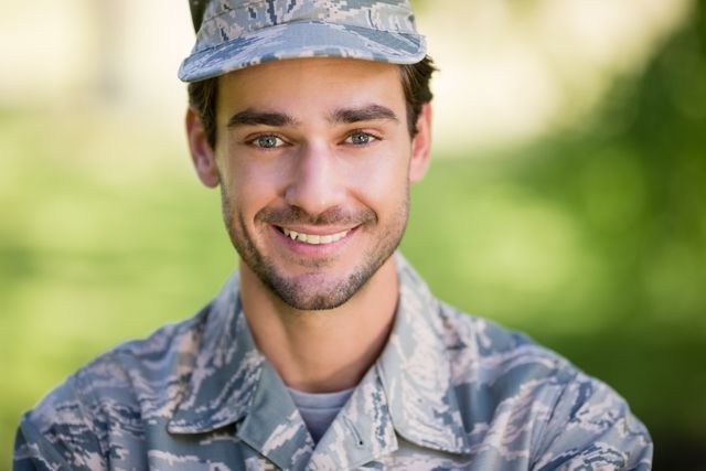 Young soldier in camouflage uniform smiling confidently in a park. Ideal for use in articles about military service, patriotism, armed forces recruitment, and personal stories of soldiers. Can also be used in promotional materials for military events or veterans' support programs.