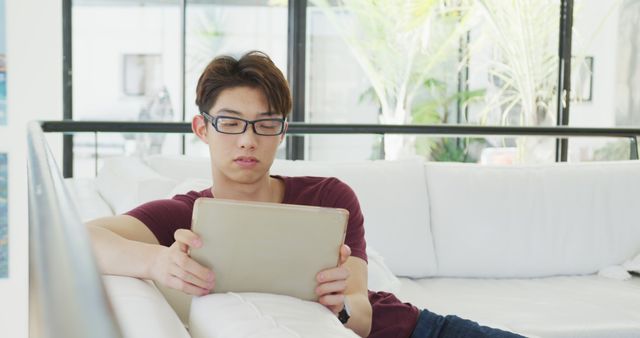 Young man wearing glasses sits casually on a white sofa at home, using a tablet. Perfect for content related to technology use, home lifestyle, relaxation, tech-savvy youth, online browsing, and indoor living spaces.