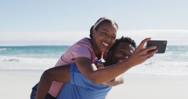 African american couple taking selfie with smartphone, man carrying woman piggyback on the beach. healthy outdoor leisure time by the sea.