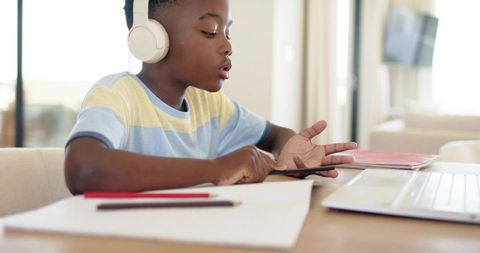 African american boy learning online at home wearing headphones using tablet and laptop