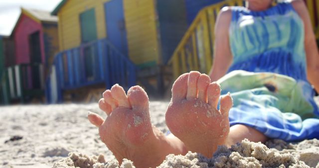 This stock photo shows a person relaxing at a sandy beach with colorful beach huts in the background. Bare feet are in the foreground, suggesting a relaxed and carefree summer vacation moment. Suitable for travel blogs, holiday promotions, leisure and relaxation themes, or advertisements for summer apparel and beach destinations.