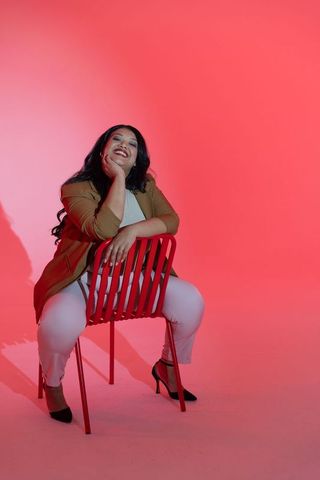Confident middle-aged woman sitting on red chair smiling showing ring wearing chic blazer