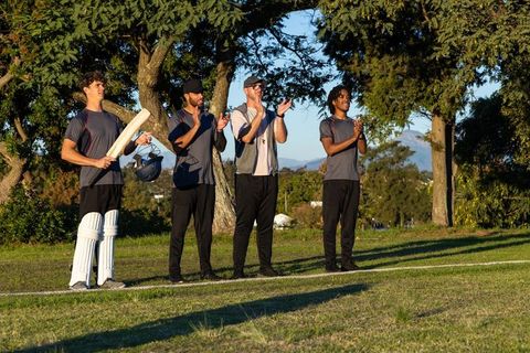 Male cricket teammates clapping on sunlit field during golden hour, batsman holding bat, pads