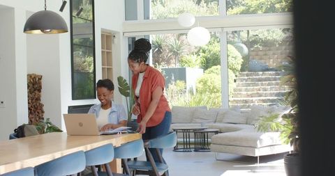 African american mother mentoring teen daughter with laptop in bright open-plan living room