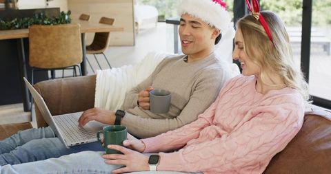 Cozy festive couple relaxing on couch with laptop, mugs, santa hat and reindeer headband