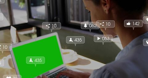 Woman typing on laptop with green screen and floating social media notifications in cafe